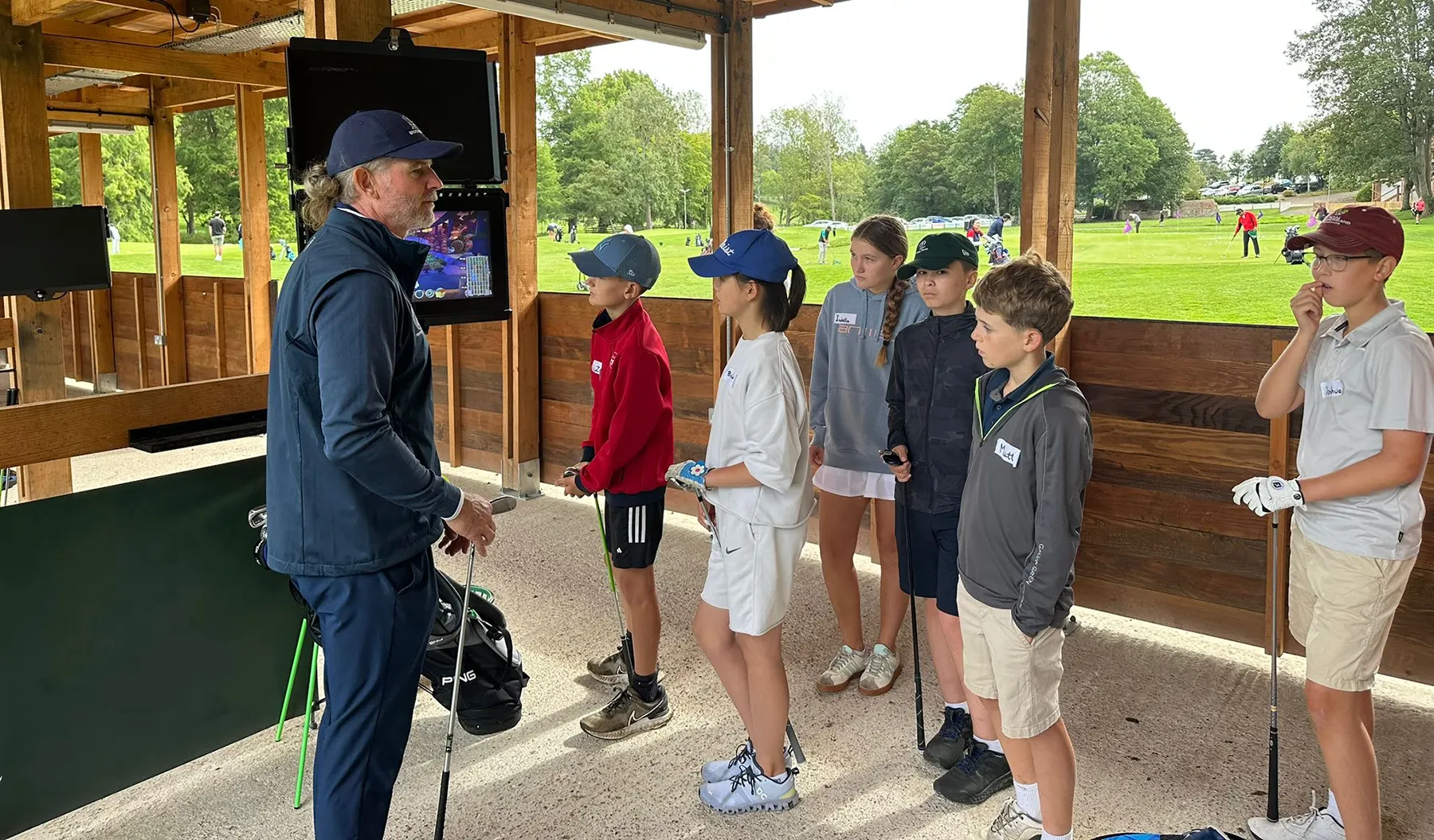 Mark Day giving a lesson to some junior golfers who listen intently at Brocket Hall's driving range. There is a Trackman screen and in the background you can see the rest of the Academy chipping area